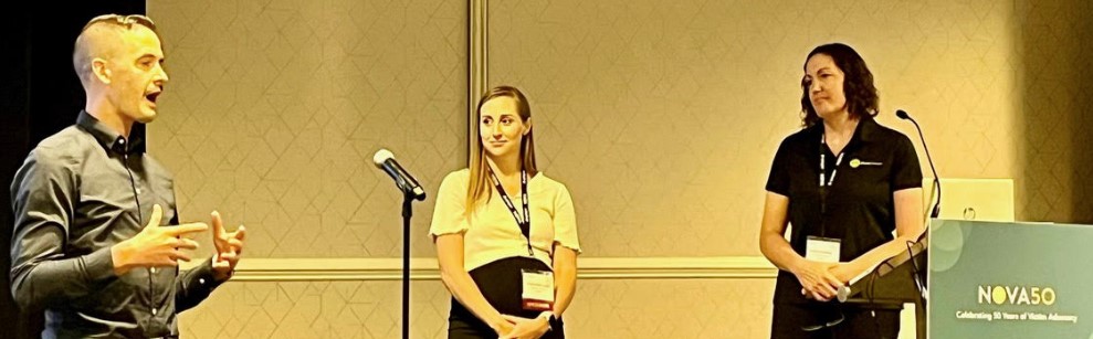 Dylan Comstock (extreme left) standing and speaking at NOVA50 Conference with two unidentified panelists.